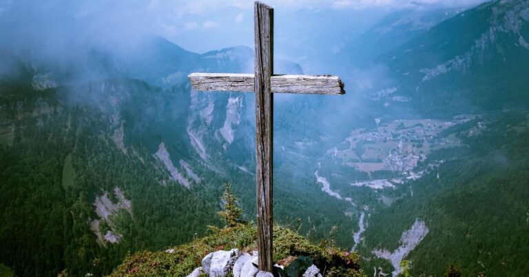 Benefits of Pleasing God - Haly Ministries (on photo: cross on hill top surrounded by mountains)