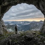 A lone figure standing at the mouth of a rocky mountain cave at dawn, with wind-blown clouds and a faint line of gold light on the horizon, conveying a quiet whisper after storm, fire and earthquake